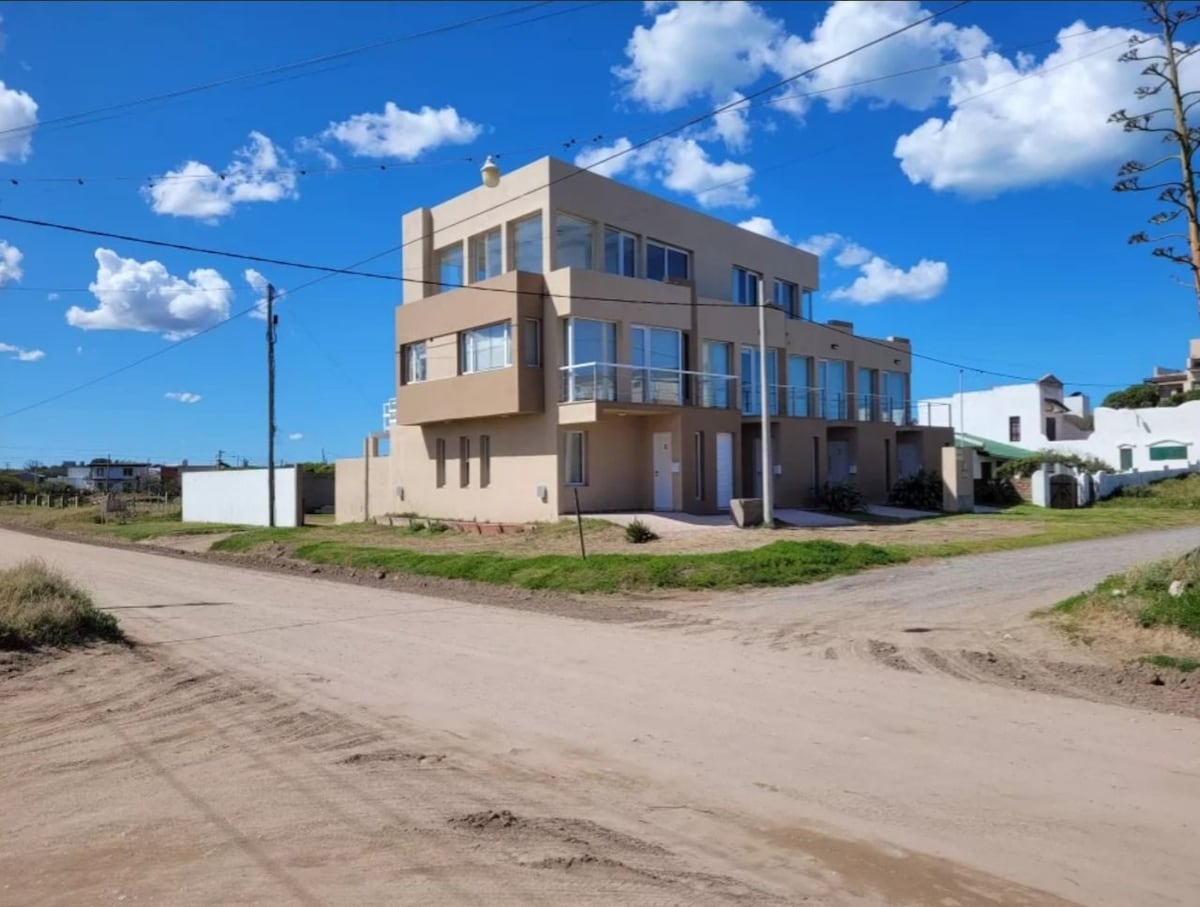 A multi-story building is shown from the street perspective, featuring large windows and balconies that provide natural light. The surrounding area includes green grass and a dirt road, with light clouds scattered in a blue sky.