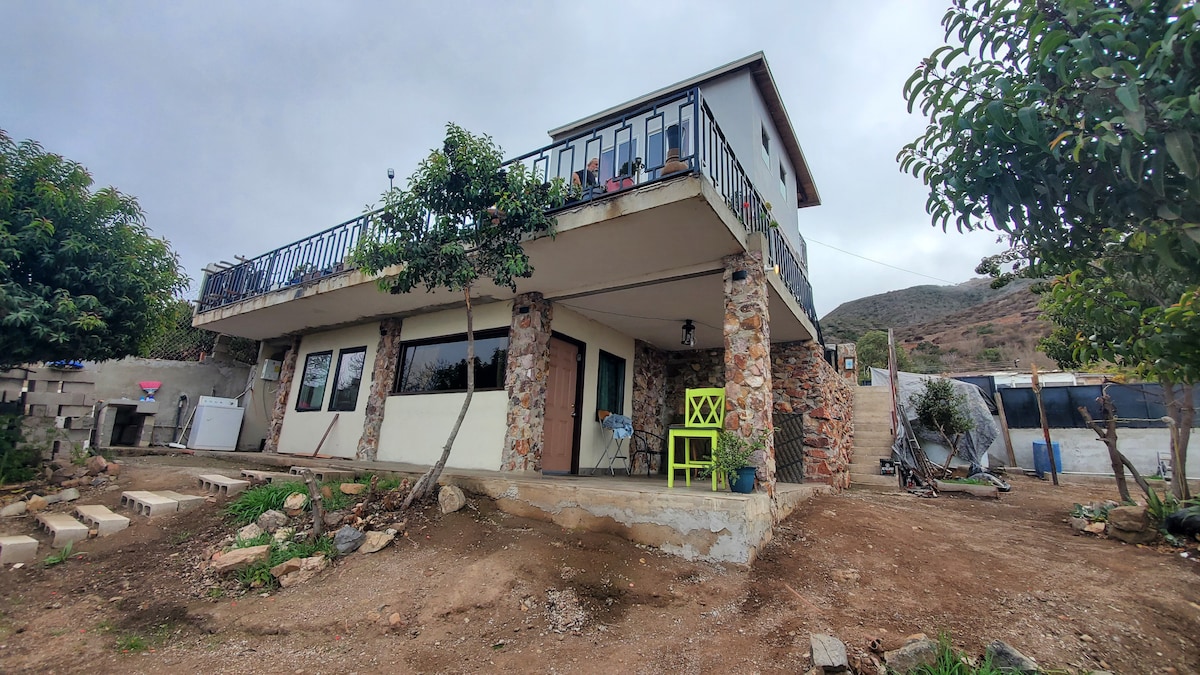A two-story building is shown, featuring a stone and concrete exterior. The upper balcony has seating and is surrounded by railings. Lush greenery is visible around the property, and a pathway leads to the front entrance, enhancing the overall welcoming presence.