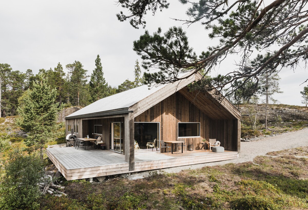A modern cabin is nestled among greenery, featuring a sloped roof and wooden exterior. Large glass doors invite natural light, and a spacious deck is partially visible, furnished with tables and chairs. The surrounding landscape includes trees and low shrubs, enhancing the natural setting.