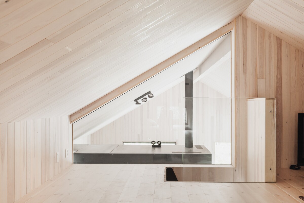 An airy loft space is showcased, with wooden paneling on the walls and ceiling. A large triangular window reflects light, creating a bright atmosphere. A minimalist shelf is seen along one wall, and ample floor space is available for relaxation or activities.
