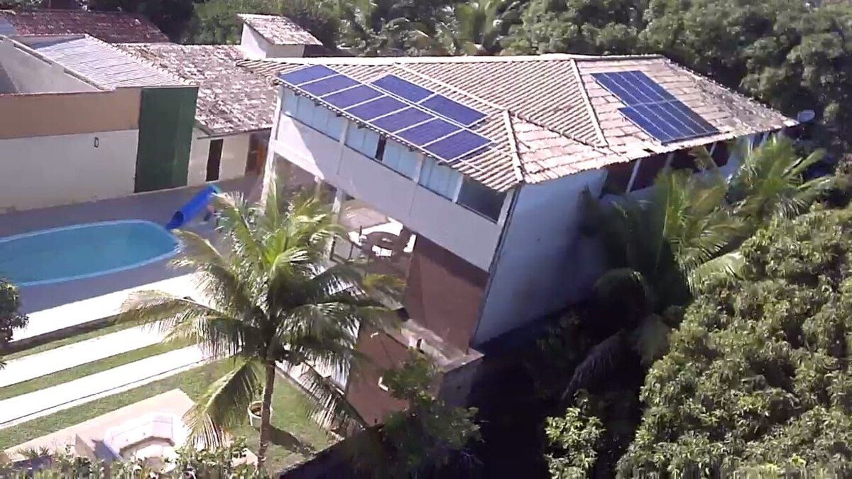 An aerial view showcases a modern house with solar panels on the sloped roof. A swimming pool is visible in the foreground, surrounded by landscaped greenery and palm trees. The structure features a mix of natural materials and open spaces, emphasizing a harmonious connection with the outdoors.