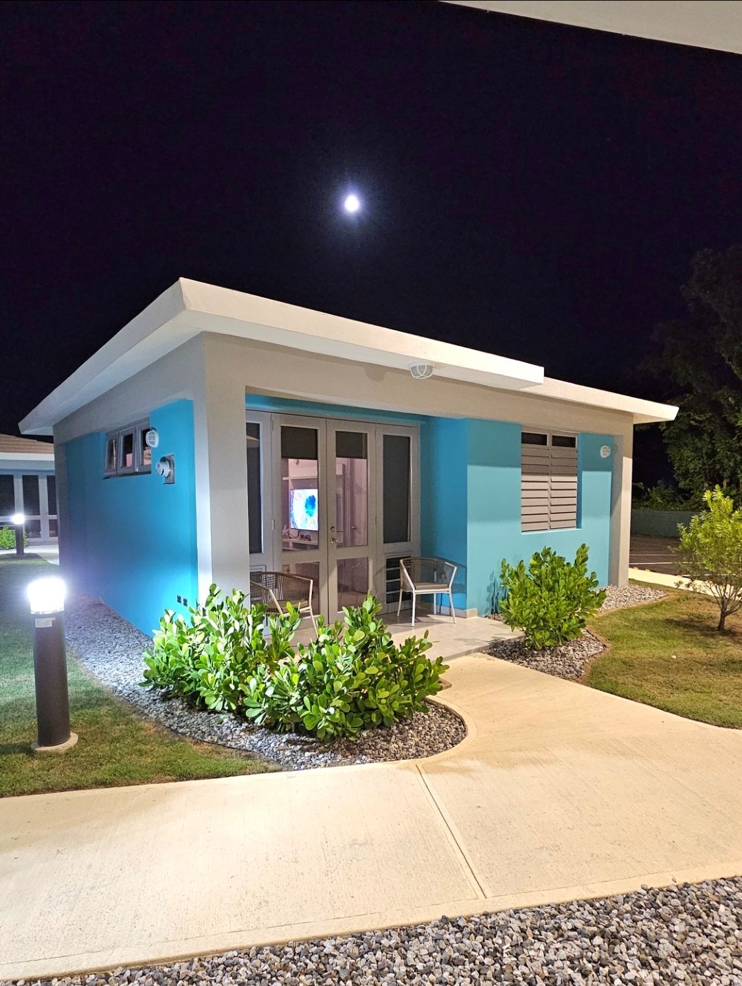 A modern villa is illuminated under the moonlight, featuring a bright blue exterior and large windows. A cozy seating area is visible on the porch, surrounded by well-maintained greenery and gravel pathways. Soft outdoor lighting enhances the inviting appearance of the property.