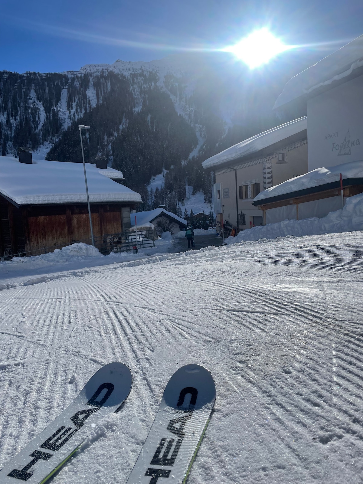 A snow-covered path is visible, lined by charming wooden chalets with steep roofs. The sun shines brightly above the surrounding mountains, casting a warm light on the scene. Ski tips from the foreground indicate an active snow sports environment.