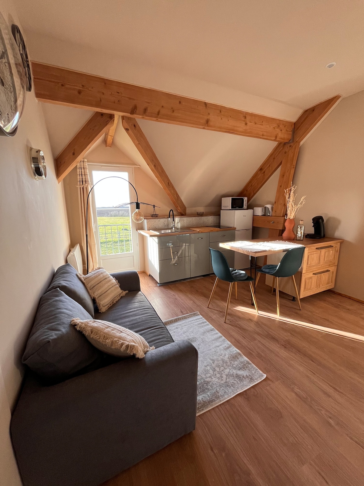 A cozy kitchenette area features wooden beams overhead, with a small dining table set for two. A comfortable gray sofa invites relaxation, while a large window offers views of the surrounding greenery. Essential appliances, including a microwave and coffee machine, complete the inviting space.