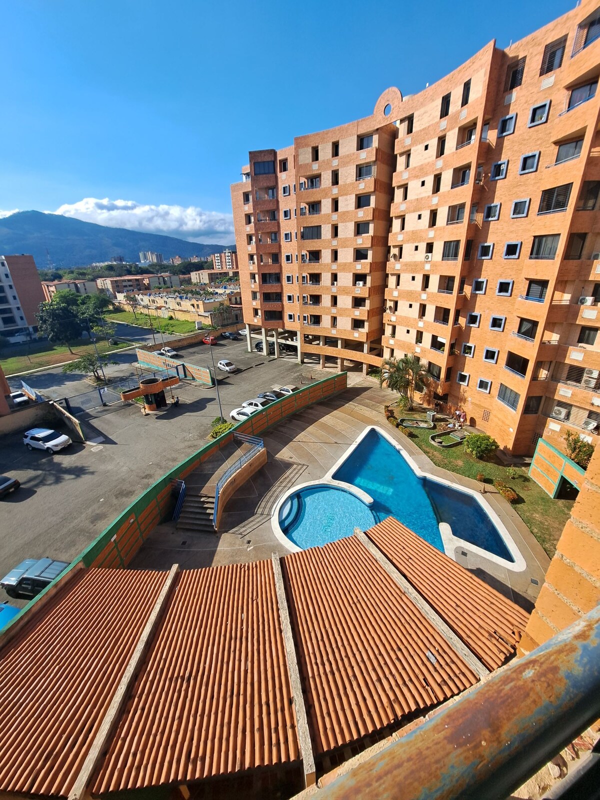 A multi-story building is viewed from above, showcasing a curved pool surrounded by a sunbathing area. Parking spaces are visible adjacent to the building, with a landscaped area adding greenery. The sky is blue, and mountains are faintly seen in the background.