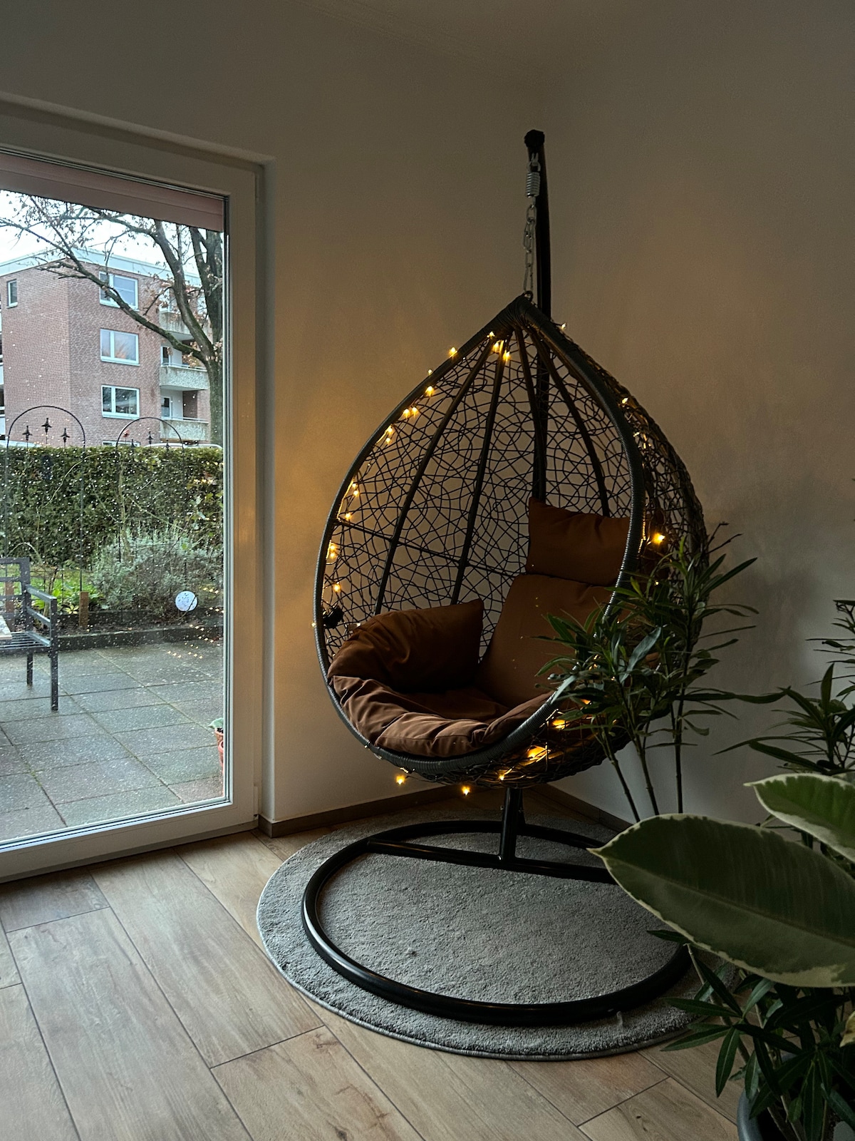 A hanging chair with soft cushions is shown, surrounded by plants and soft lighting. Large glass doors allow natural light to brighten the space, leading to an outdoor area. Warm, inviting tones create a cozy atmosphere in this corner of the room.