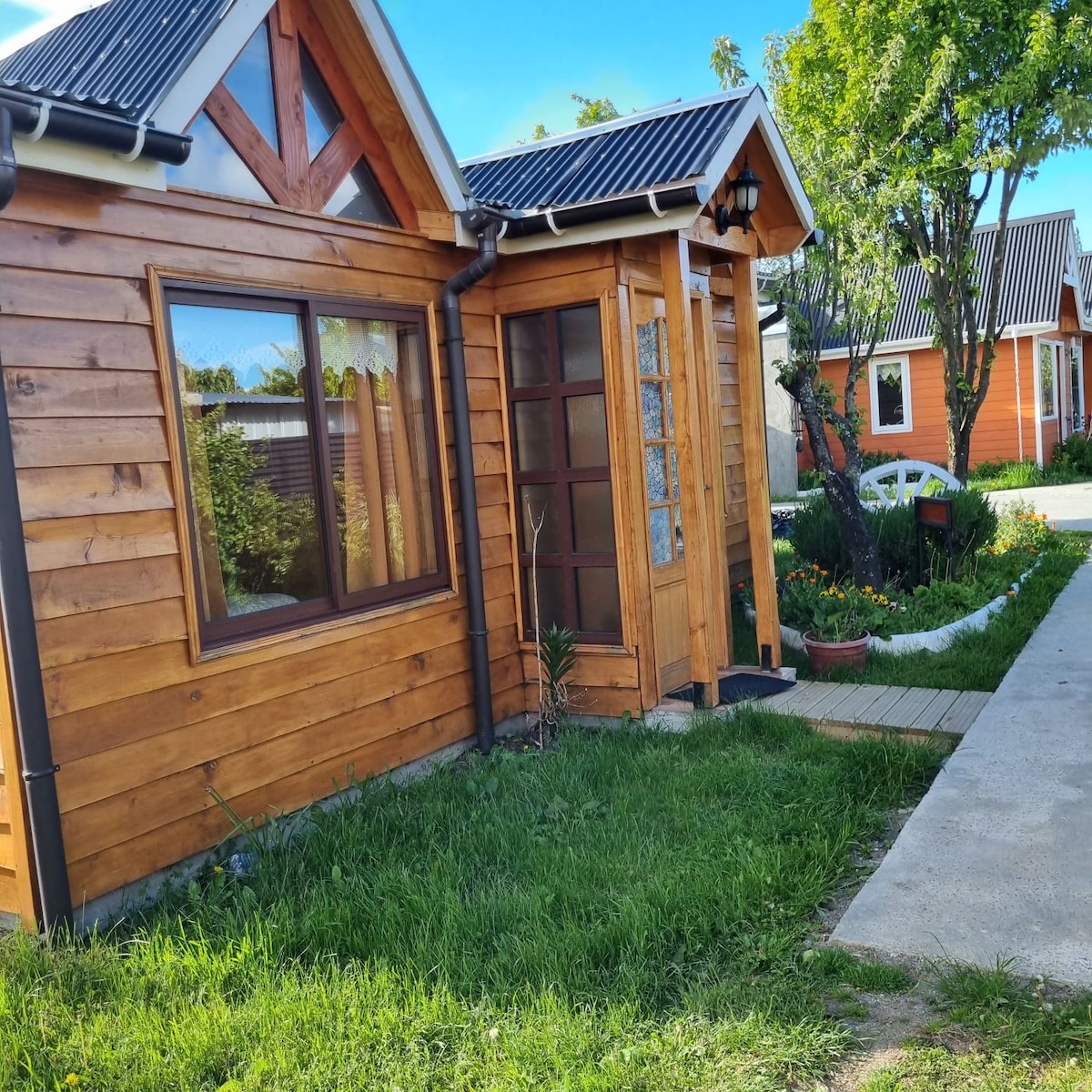 A charming wooden exterior showcases a welcoming entrance with a glass door. Lush green grass surrounds the path leading to the house, and colorful flowers add a touch of vibrancy to the landscape. The sloped roof and gabled design highlight the home's cozy architecture.