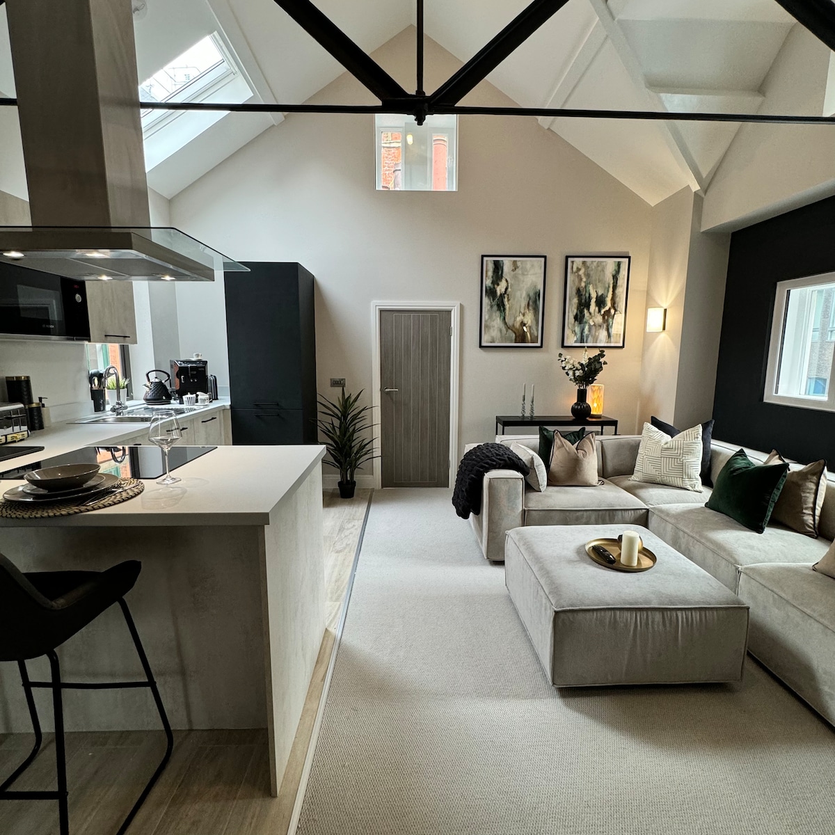 A contemporary living area features a spacious sectional sofa adorned with accent pillows. A stylish coffee table sits in front, with decorative candles arranged on top. The kitchen area is visible, showcasing modern appliances and a sleek design, complemented by natural light from the skylight.