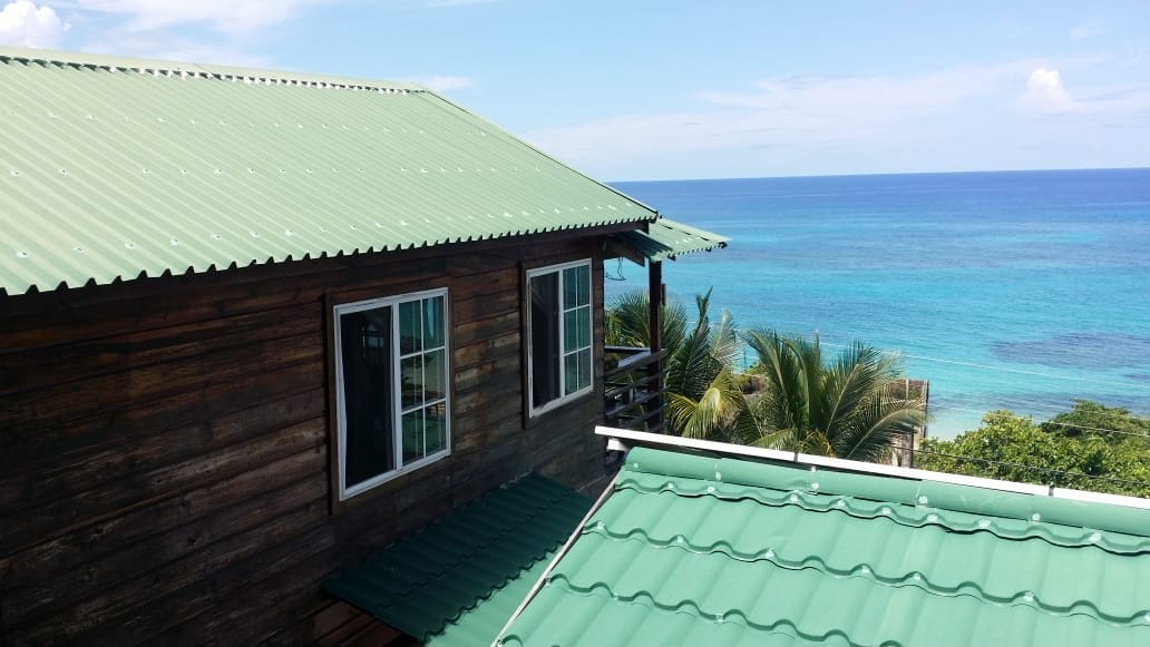 A wooden bungalow is displayed against a backdrop of vibrant turquoise waters. The structure features a green metal roof and white-framed windows, with palm trees visible in the distance, emphasizing the coastal setting.