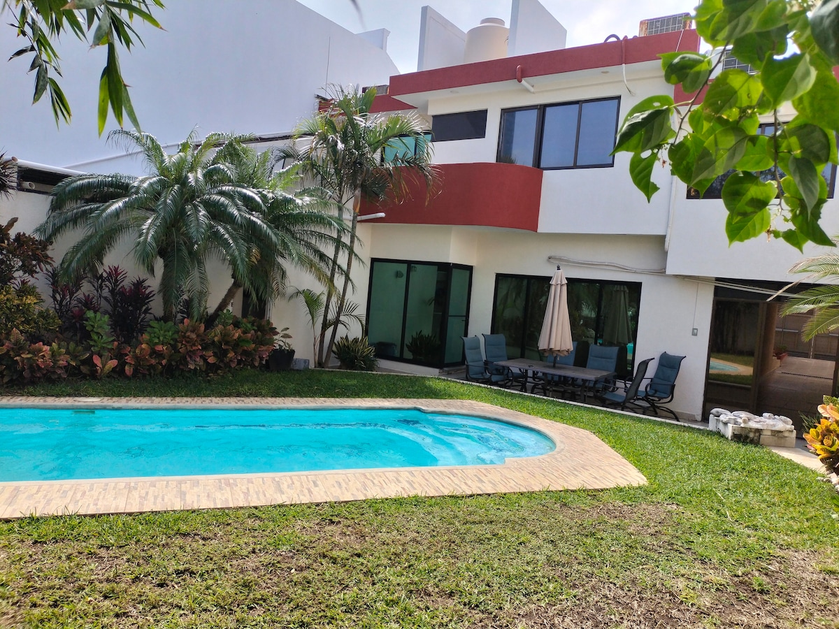 A view of the private pool surrounded by lush greenery and colorful plants. Comfortable outdoor seating and a dining area are visible under a shaded umbrella. The house features large glass doors that allow natural light to enter the living space.