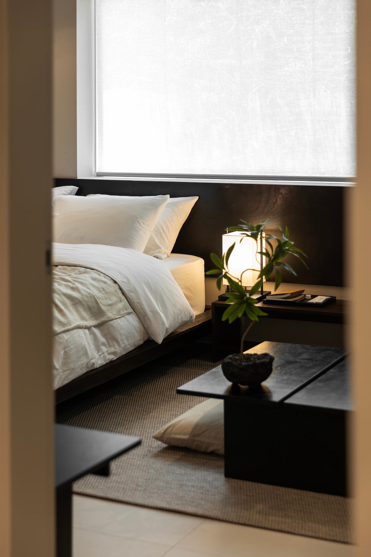 A serene bedroom features a spacious bed adorned with white linens and accent pillows. A minimalist black nightstand holds a softly glowing lamp. A small potted plant adds a touch of greenery, while natural light filters through the sheer window coverings.