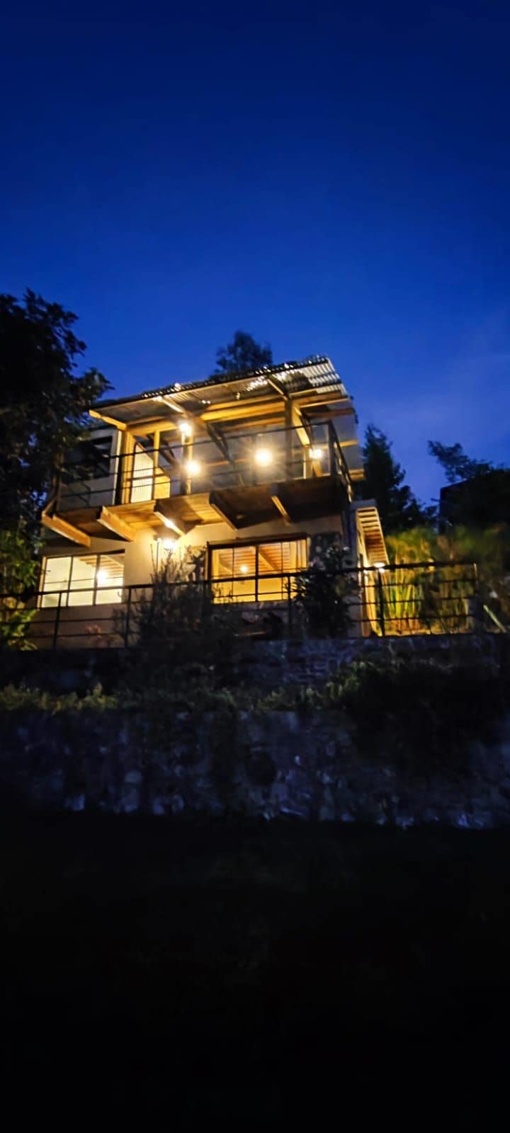 A modern multi-level house is illuminated at night, showcasing large glass windows and wooden balconies. The surrounding greenery adds to the serene ambiance, while soft lighting emphasizes the structural design against the darkening sky.