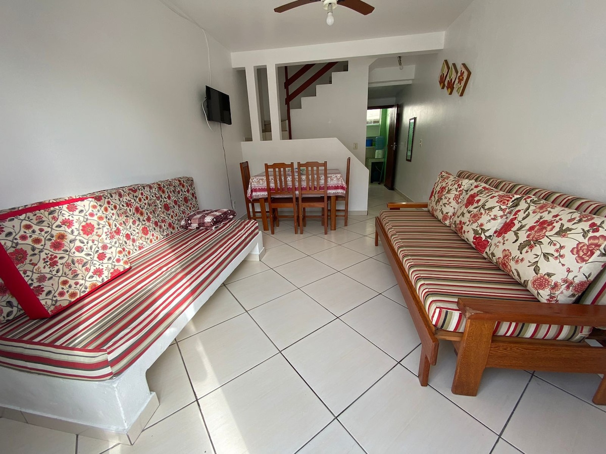 A living area is presented with two patterned sofas, one with floral design and the other with striped fabric. A wooden dining table with matching chairs is positioned nearby. Light-colored tiles cover the floor, and a ceiling fan is mounted above.