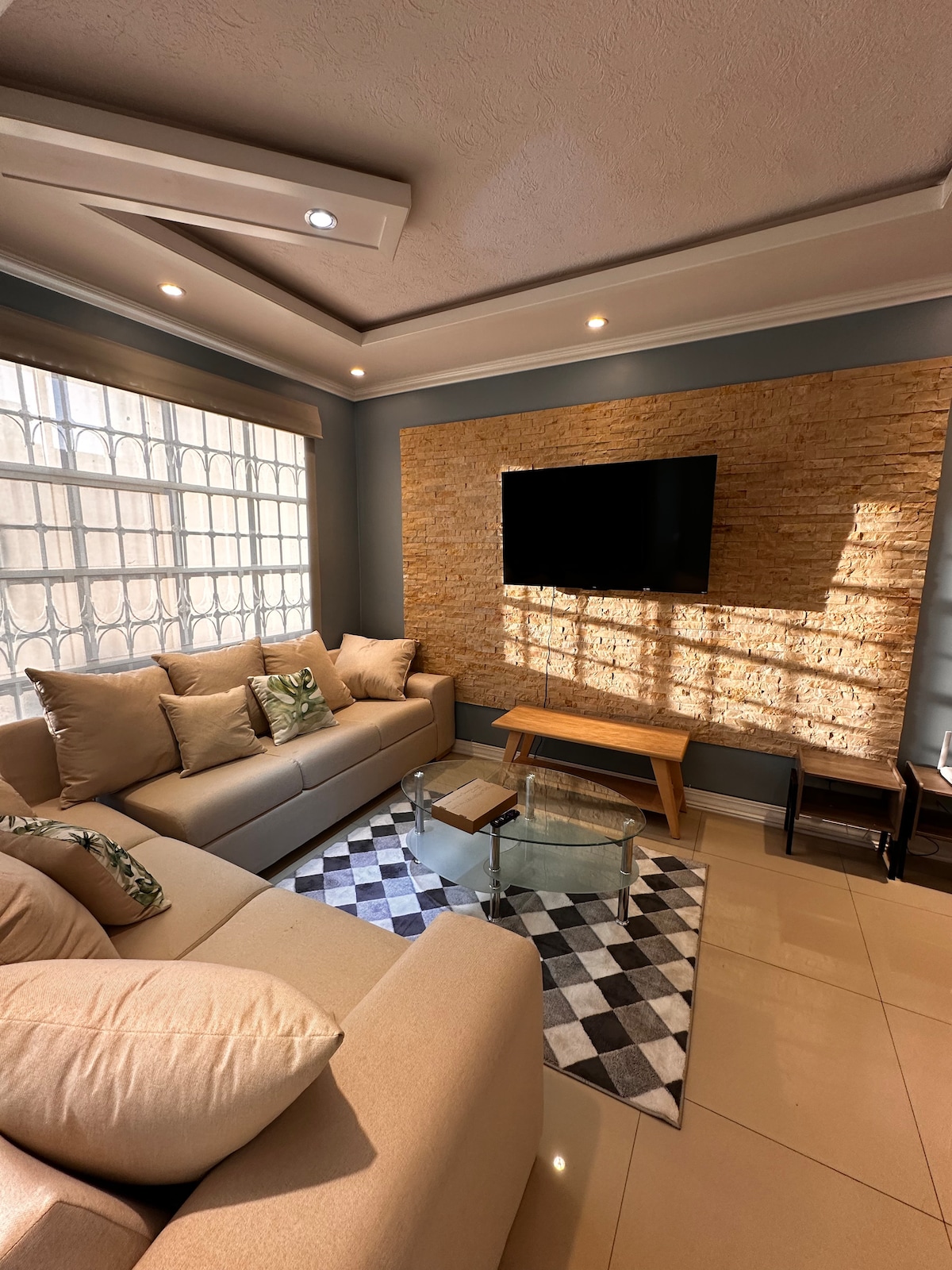 A comfortable living area features a soft beige sofa adorned with pillows. A patterned rug lies beneath a glass coffee table. The wall is accented with natural stone, and a flat-screen TV is mounted above a wooden console, illuminated by natural light from large windows.