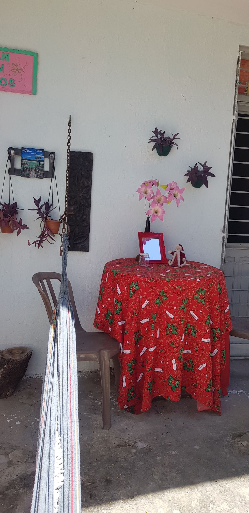 A small outdoor seating area is presented with a round table draped in a vibrant red tablecloth adorned with festive patterns. A single chair is positioned beside the table, while potted plants hang on the wall, adding a touch of greenery to the space.