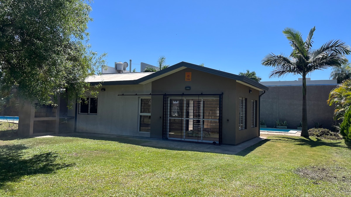 The exterior of a modern house is captured under clear blue skies. The house features a spacious lawn with well-maintained grass, framed by palm trees. Large windows, secured by modern grilles, allow natural light into the interior while offering a view of the outdoor space.