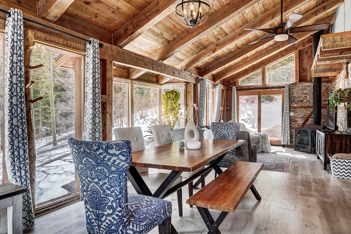 A spacious dining area features a rustic wood table accompanied by a bench and upholstered chairs. Large windows allow natural light to illuminate the wooden beams and walls. The cozy ambiance is enhanced by a ceiling fan and decorative accents throughout the room.