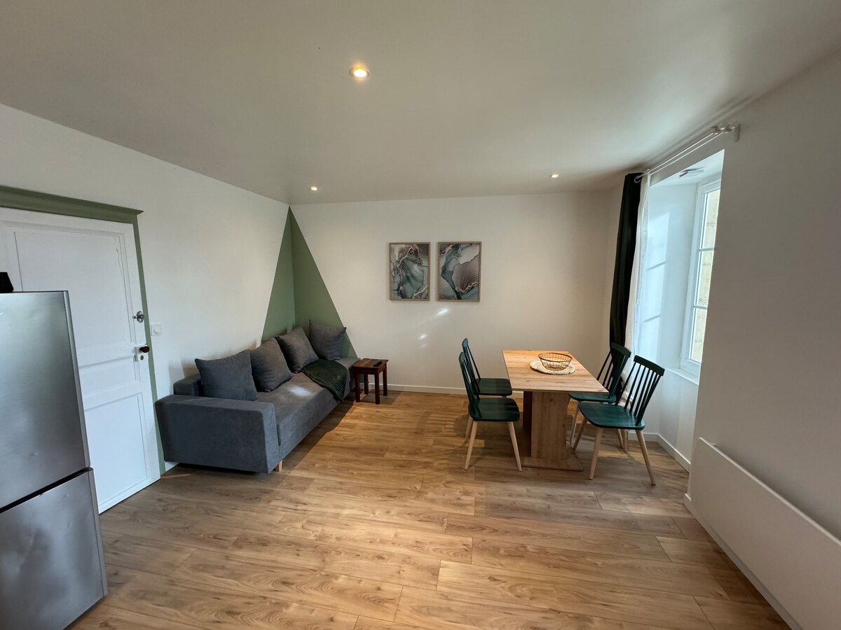 The living area features a comfortable gray sofa, paired with a wooden dining table and green chairs. Two pieces of wall art adorn the walls, while natural light filters through the window, accentuating the wooden floor and brightening the space.