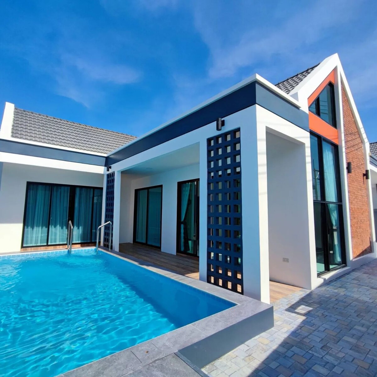 A modern villa is positioned beside a clear blue pool, with large windows allowing plenty of natural light. The architectural design includes a combination of brick and smooth white walls, complemented by contrasting dark accents.