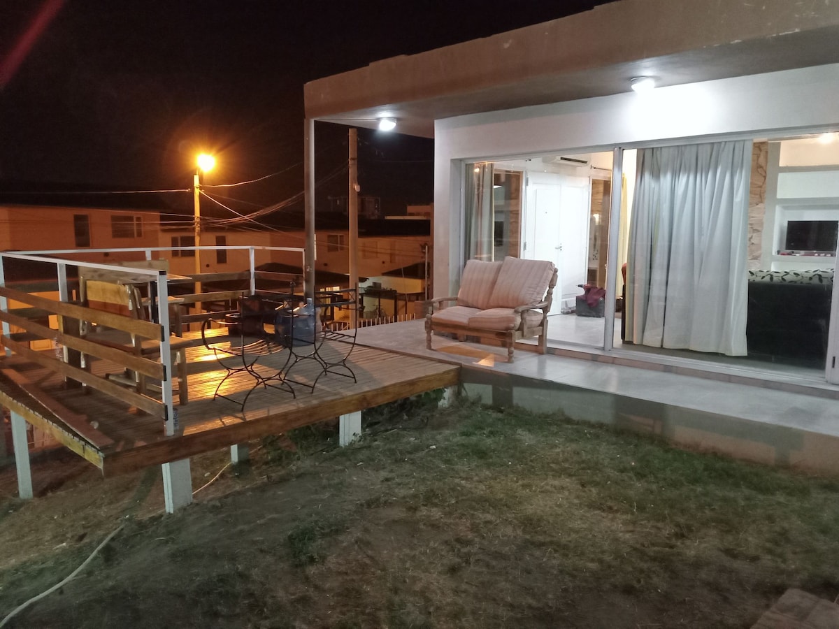 An outdoor deck features a wooden railing and a comfortable seating area, illuminated by nearby lamps. The adjacent indoor space is visible through large glass doors, showcasing soft drapes and a cozy interior setting.