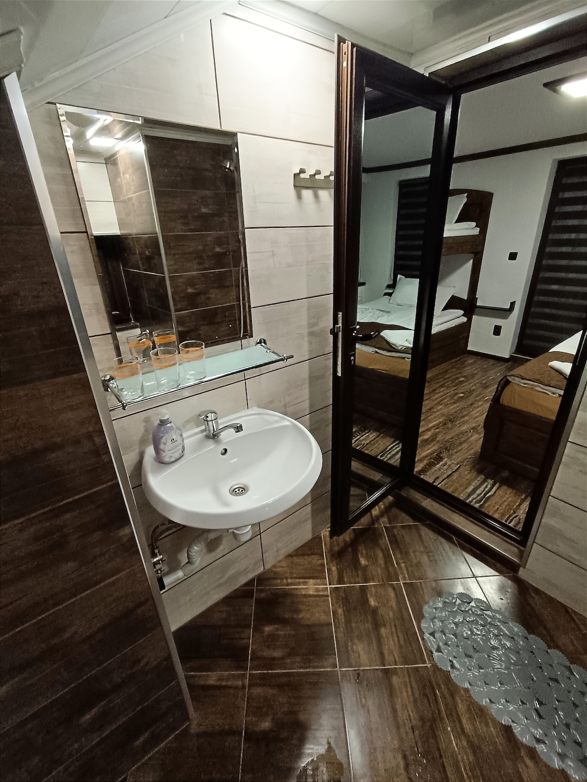 The bathroom features modern fixtures, including a sink with a mirrored shelf above and a spacious shower area. Dark tile flooring contrasts with light wall tiles, creating a clean and functional space. A glimpse of the bedroom can be seen through the glass door.