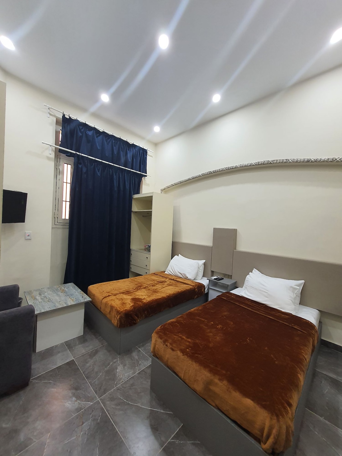 A well-lit room features two separate beds covered with soft brown blankets. Tall windows are adorned with dark curtains. A small chair and table are positioned in a corner, while a flat-screen TV is mounted on the wall, providing modern amenities.