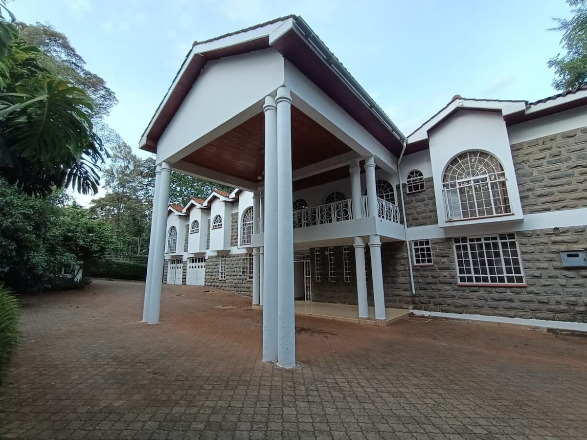 The exterior of a spacious house is observed, featuring a large covered porch supported by four columns. The facade is made of stone, accented by arched windows with decorative grills. A paved driveway extends toward the entrance, surrounded by lush greenery.