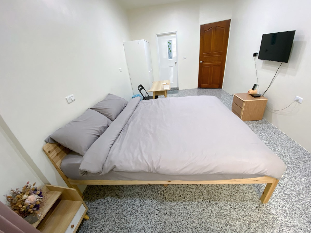A clean and comfortable space is shown with a spacious bed featuring gray bedding. A wooden table and chair are positioned near a wall, while a small bedside table holds a decorative vase. A television is mounted on the wall opposite the bed.