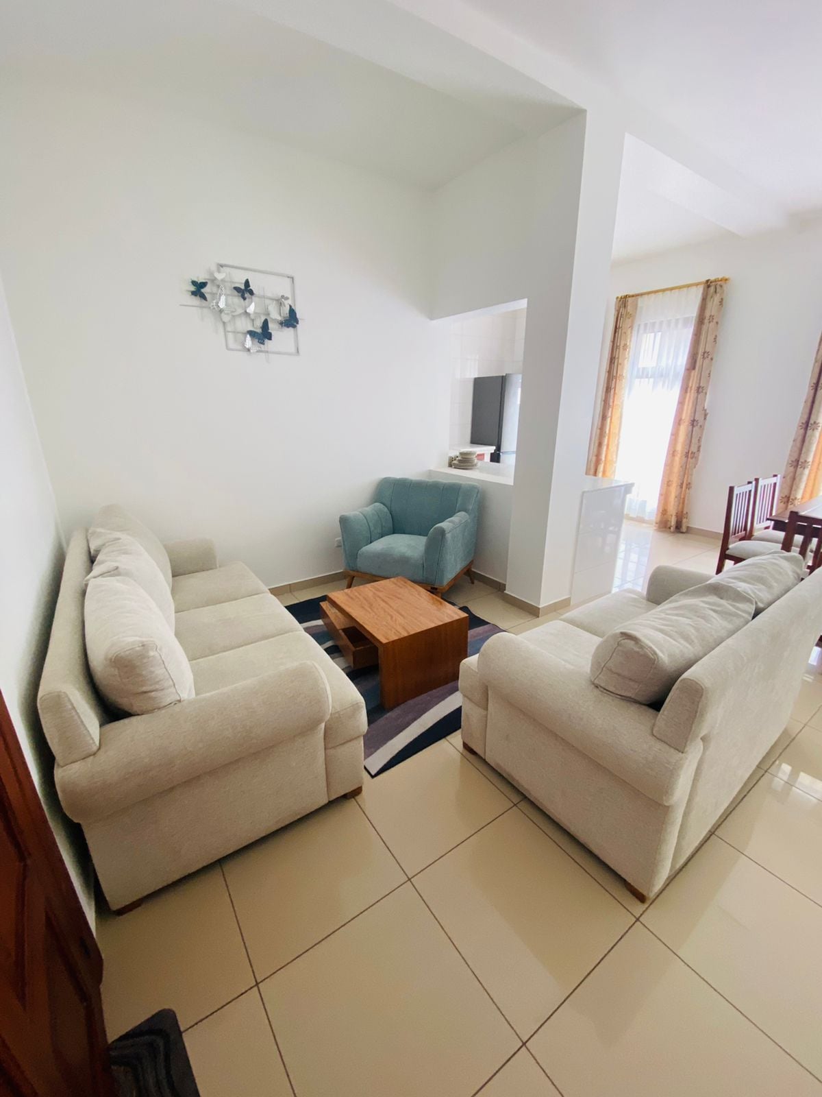 A cozy seating area features two light-colored sofas positioned across from a small wooden coffee table. A single, blue accent chair adds a pop of color. Natural light enters from a nearby window, complemented by curtains that softly frame the space.