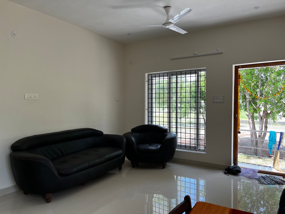 A spacious living area is presented with a sleek black sofa and a matching armchair. A large window allows natural light to fill the room, revealing views of the surrounding greenery. Light-colored walls and polished floors enhance the open atmosphere.