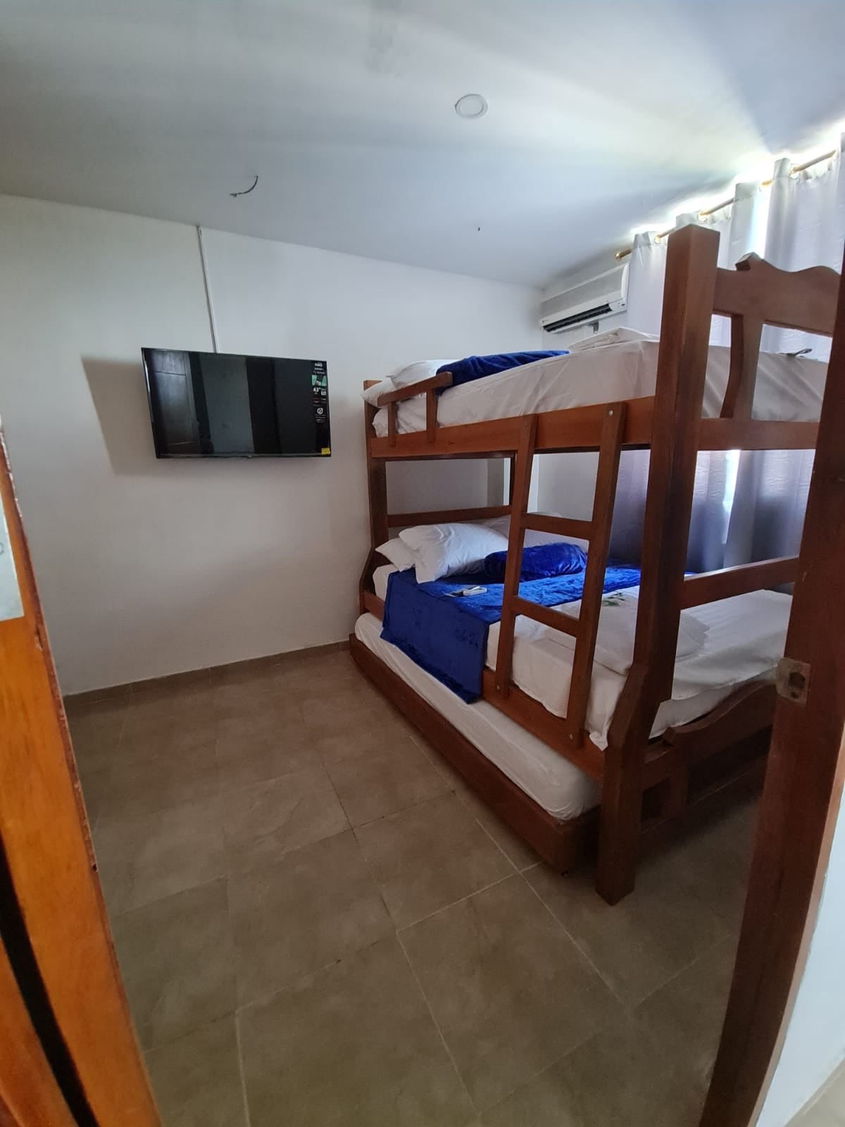 A cozy sleeping area is depicted featuring a wooden bunk bed with two levels, each presenting neatly arranged white bedding accompanied by blue throws. A flat-screen television is mounted on the wall, and natural light is seen filtering through partially closed curtains.