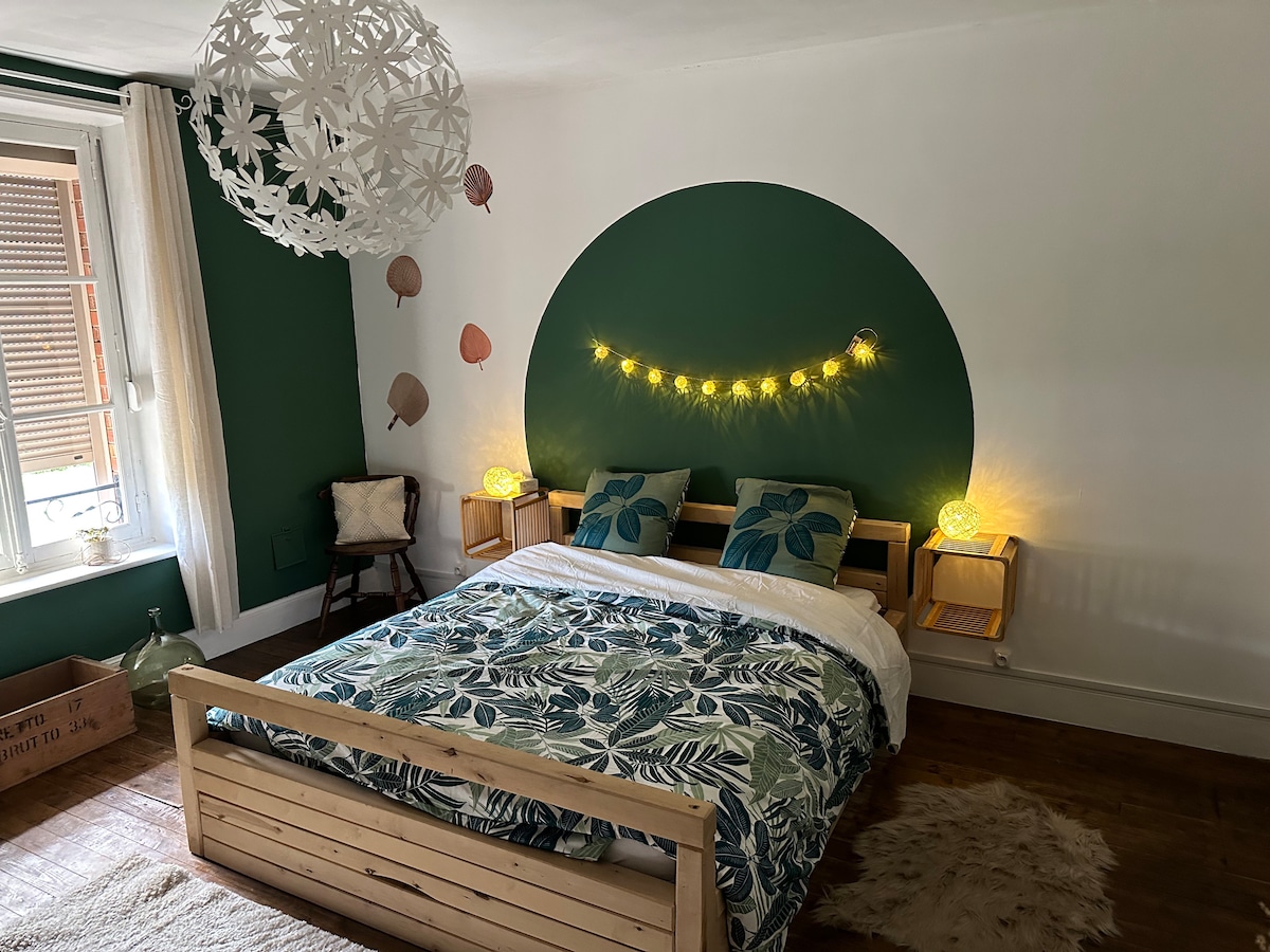A cozy bedroom features a wooden bed adorned with a leafy patterned duvet and white linens. Soft lighting is provided by decorative bulbs on the wall and bedside tables. Green walls complement wall art, while natural light filters through a window with wooden shutters.