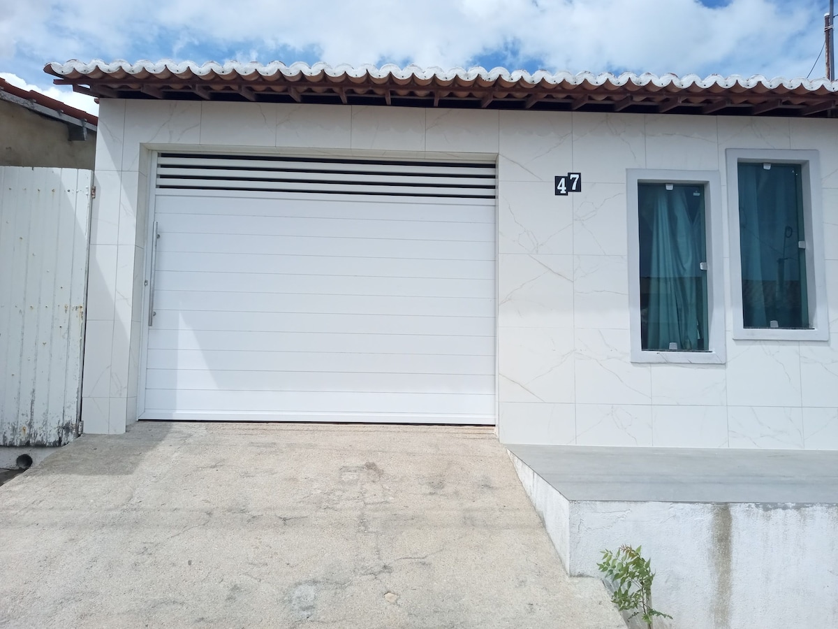 A modern exterior features a smooth white facade with light marble patterns. A large garage door is visible, alongside two windows dressed in sheer curtains. The entrance is approached by a concrete walkway, leading to a small raised platform.