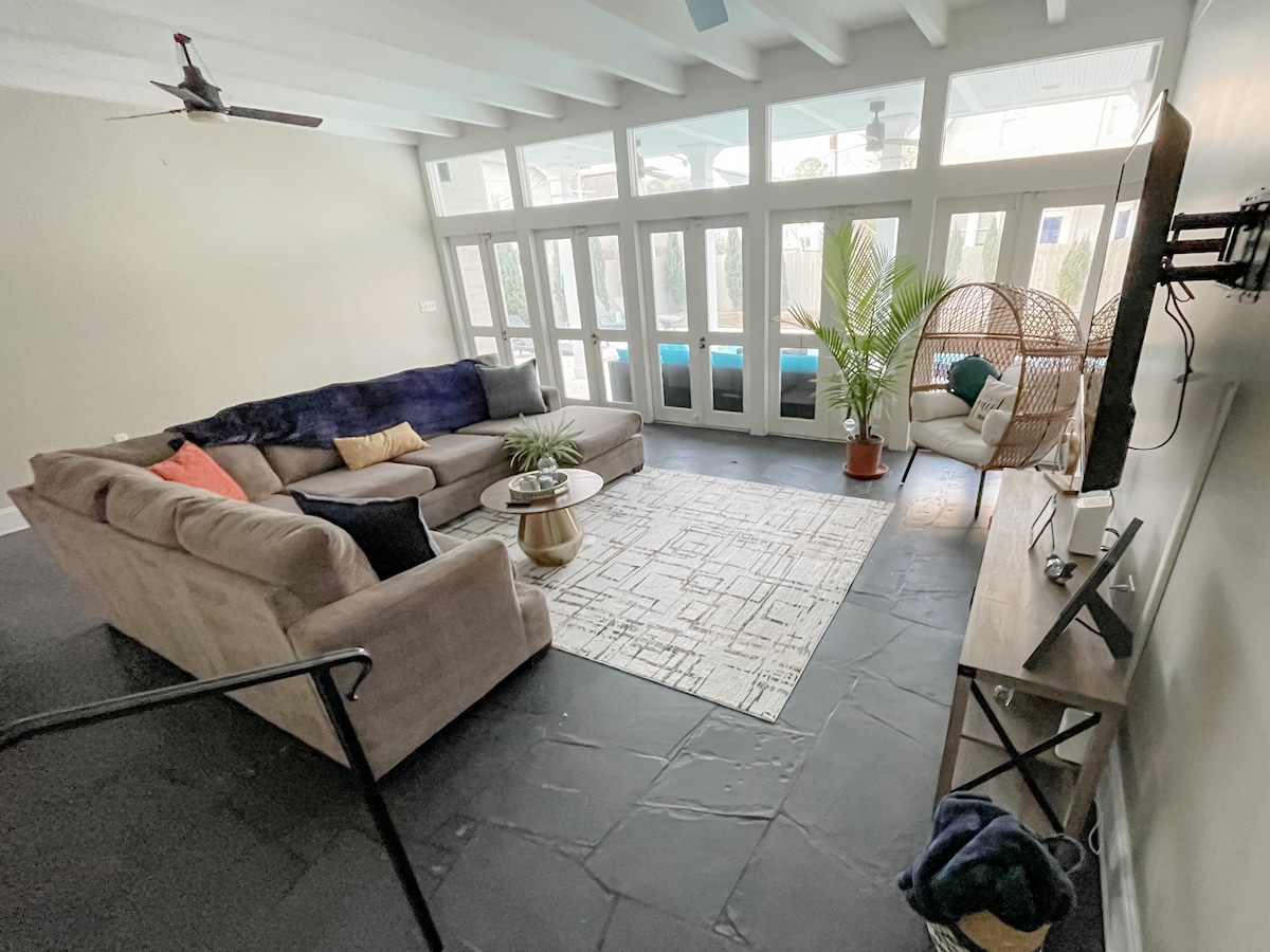 A spacious sitting room is showcased with a comfortable sectional sofa and a cozy armchair. Natural light fills the room through large glass doors. A small coffee table is placed on a textured area rug, while a tall plant adds a touch of greenery.