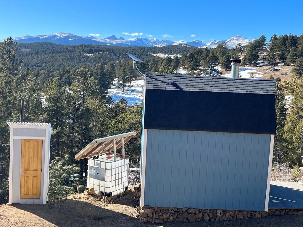 The image features a small, insulated cabin alongside a simple outhouse. The cabin displays a pitched roof with solar panels, while a water storage unit is positioned nearby. The backdrop showcases a stunning mountain landscape, with trees visible in the foreground and snow-capped peaks in the distance.