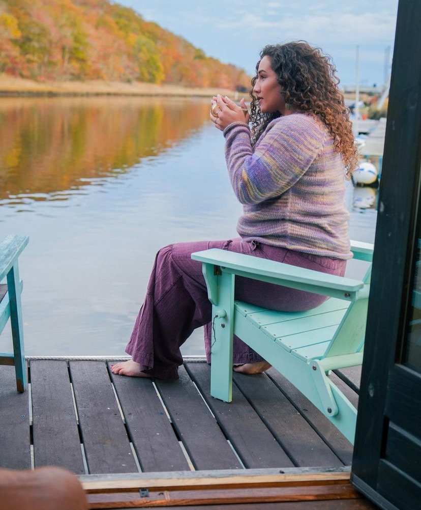A person is seated on a pastel-colored Adirondack chair on a wooden deck, facing the calm river. The tranquil water reflects the vibrant autumn foliage, while the sky is dotted with soft clouds. The serene setting emphasizes a peaceful waterfront experience.