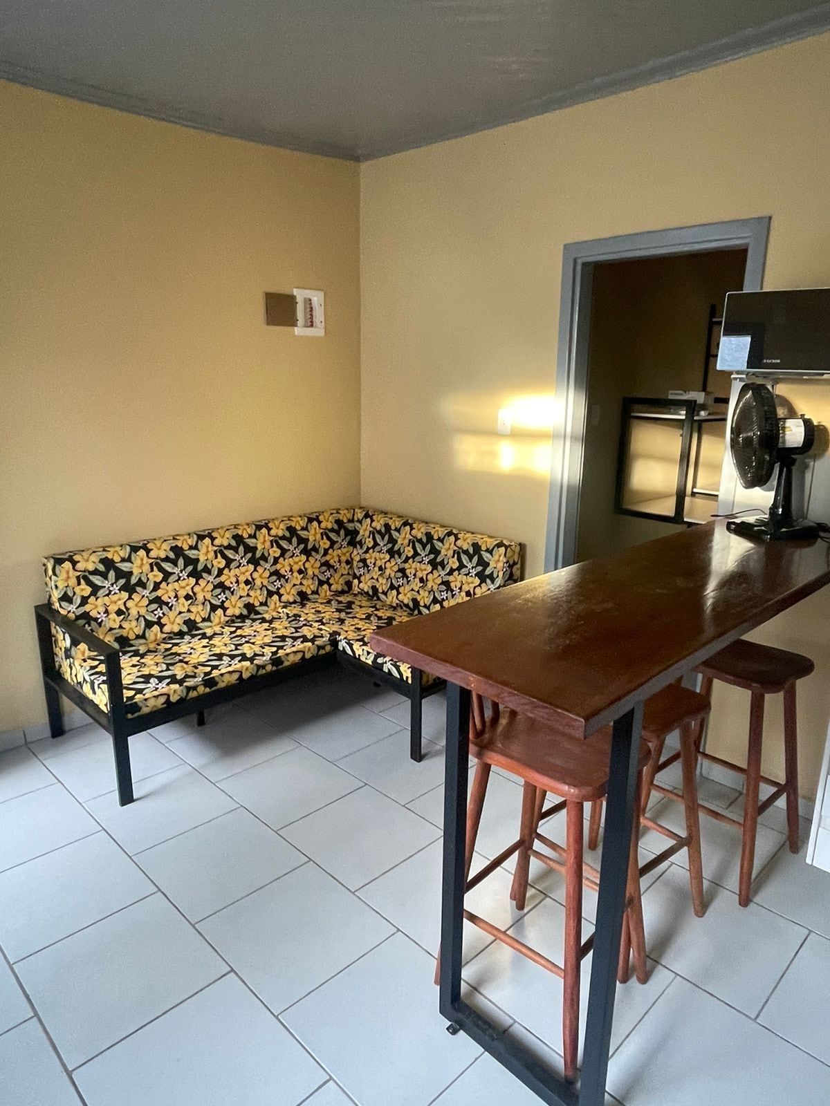 A comfortable seating area features a floral-patterned couch against a warm yellow wall. A wooden bar table with three stools is placed nearby, complementing the simple and functional design. The space is well-lit by natural light entering from a window, highlighting its clean tile flooring.