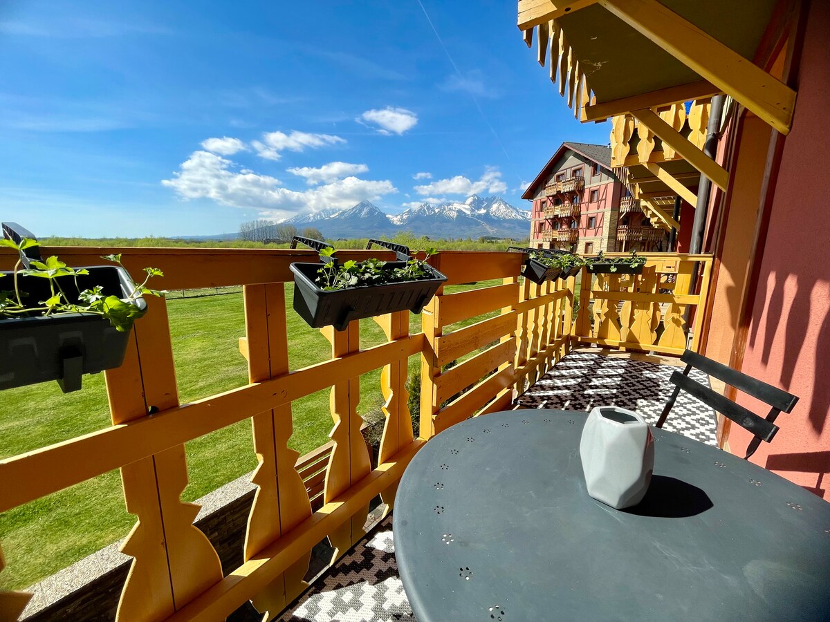 A balcony offers a panoramic view of the Tatra mountains, adorned with flower boxes. A circular table and a decorative vase occupy the space, complemented by a sunny sky and scattered clouds.