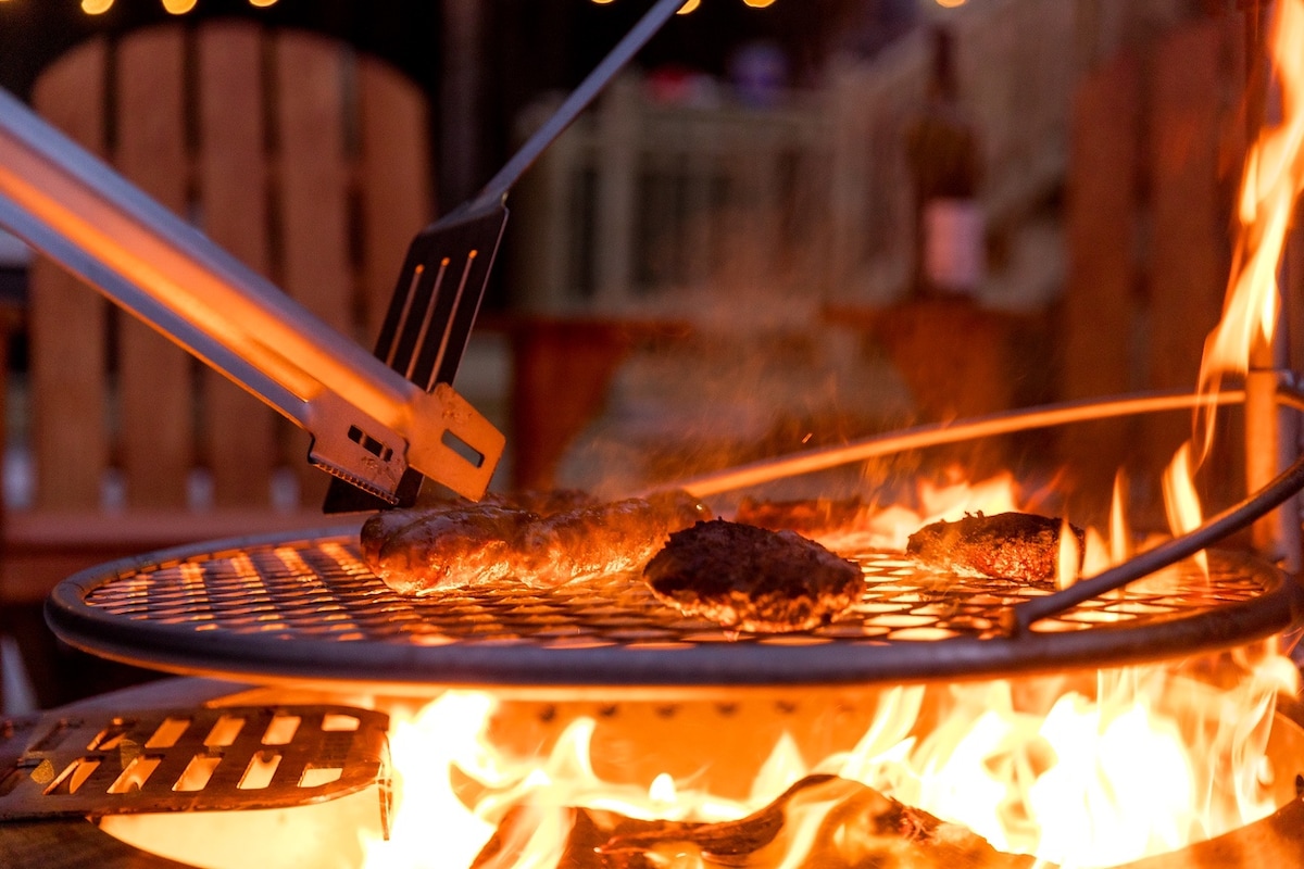 A smokeless fire pit displays grill marks on several pieces of meat, with flames dancing around them. Long grilling tongs hold one item above the grill. Warm lighting illuminates the surrounding area, enhancing the outdoor cooking experience.