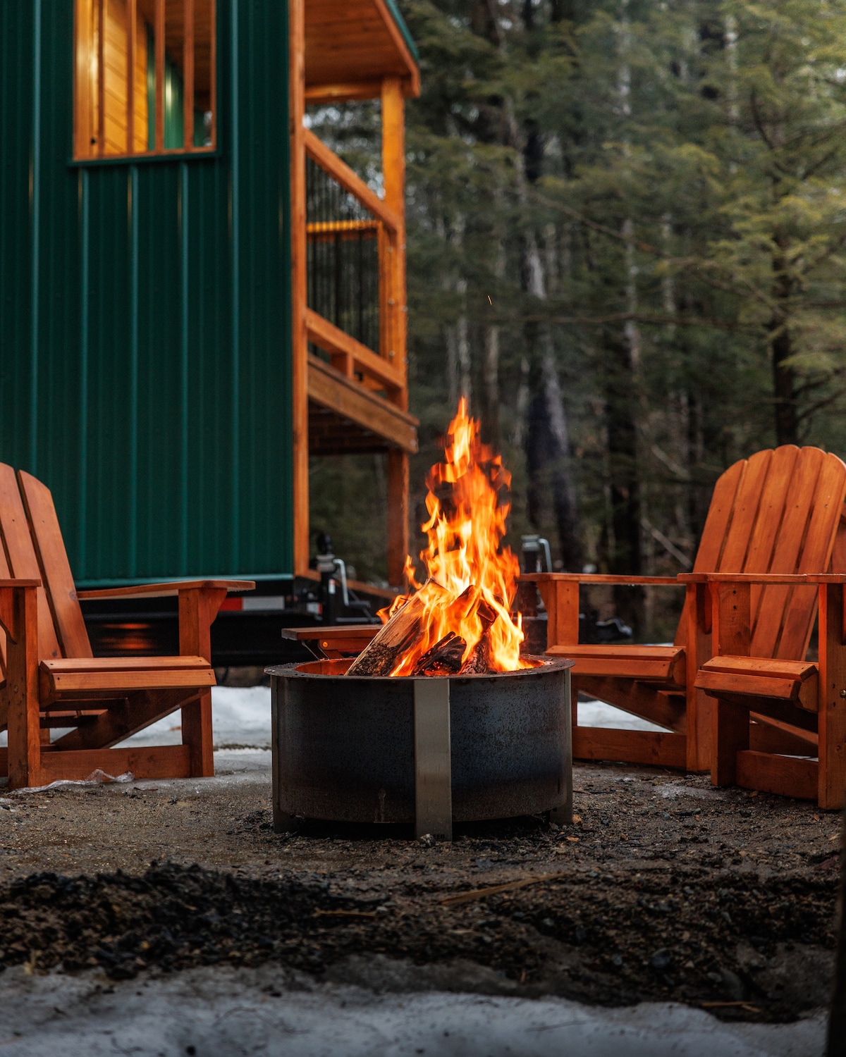 A cozy fire pit surrounded by two wooden adirondack chairs is depicted, with flames brightly illuminating the area. The backdrop features the wooden balcony of a tiny home, amidst a natural setting with trees visible in the distance.