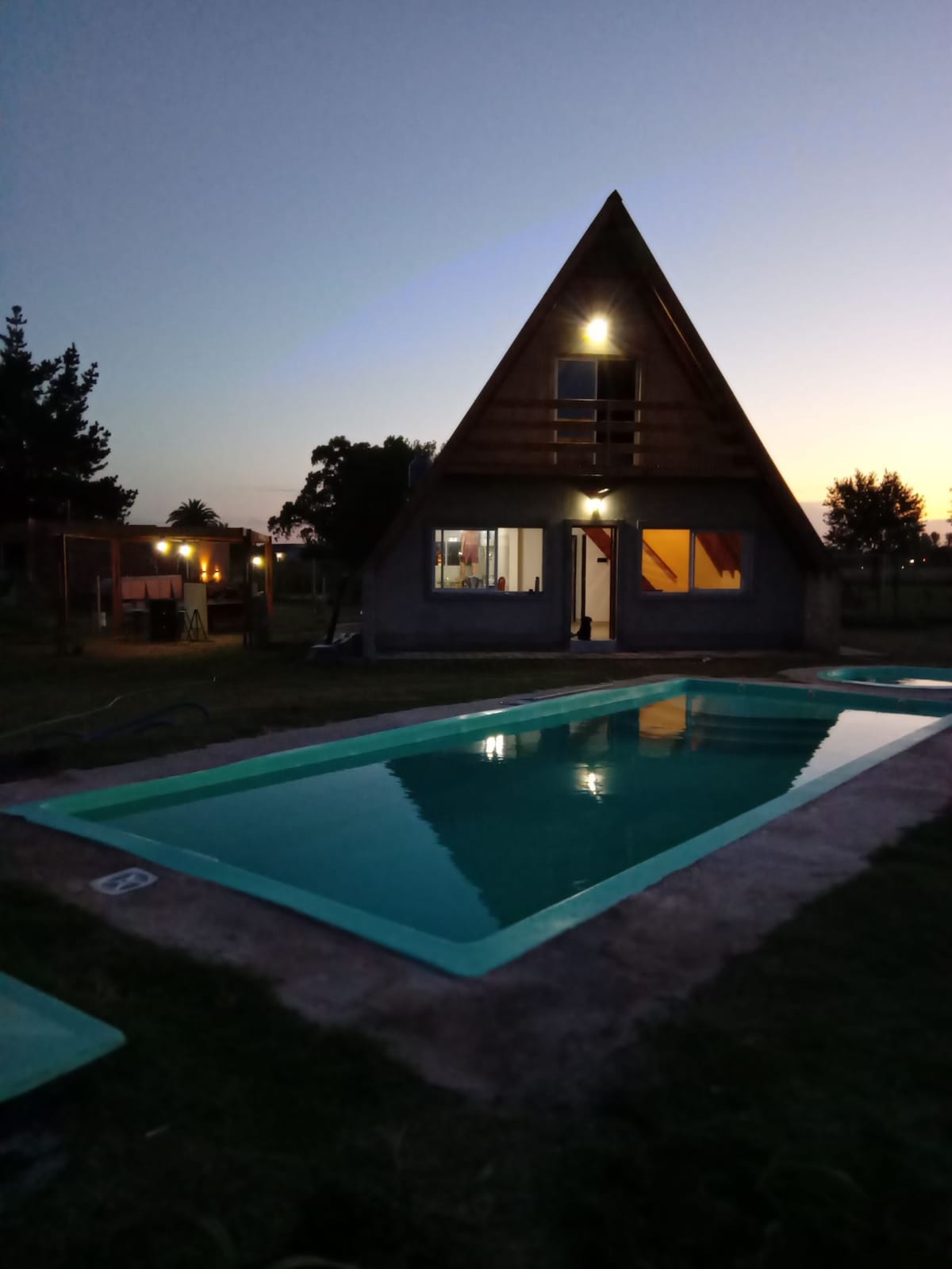 A modern a-frame house is set against a twilight sky, with a warm glow emanating from its windows. A rectangular pool is visible in the foreground, surrounded by a grassy area and trees in the background.