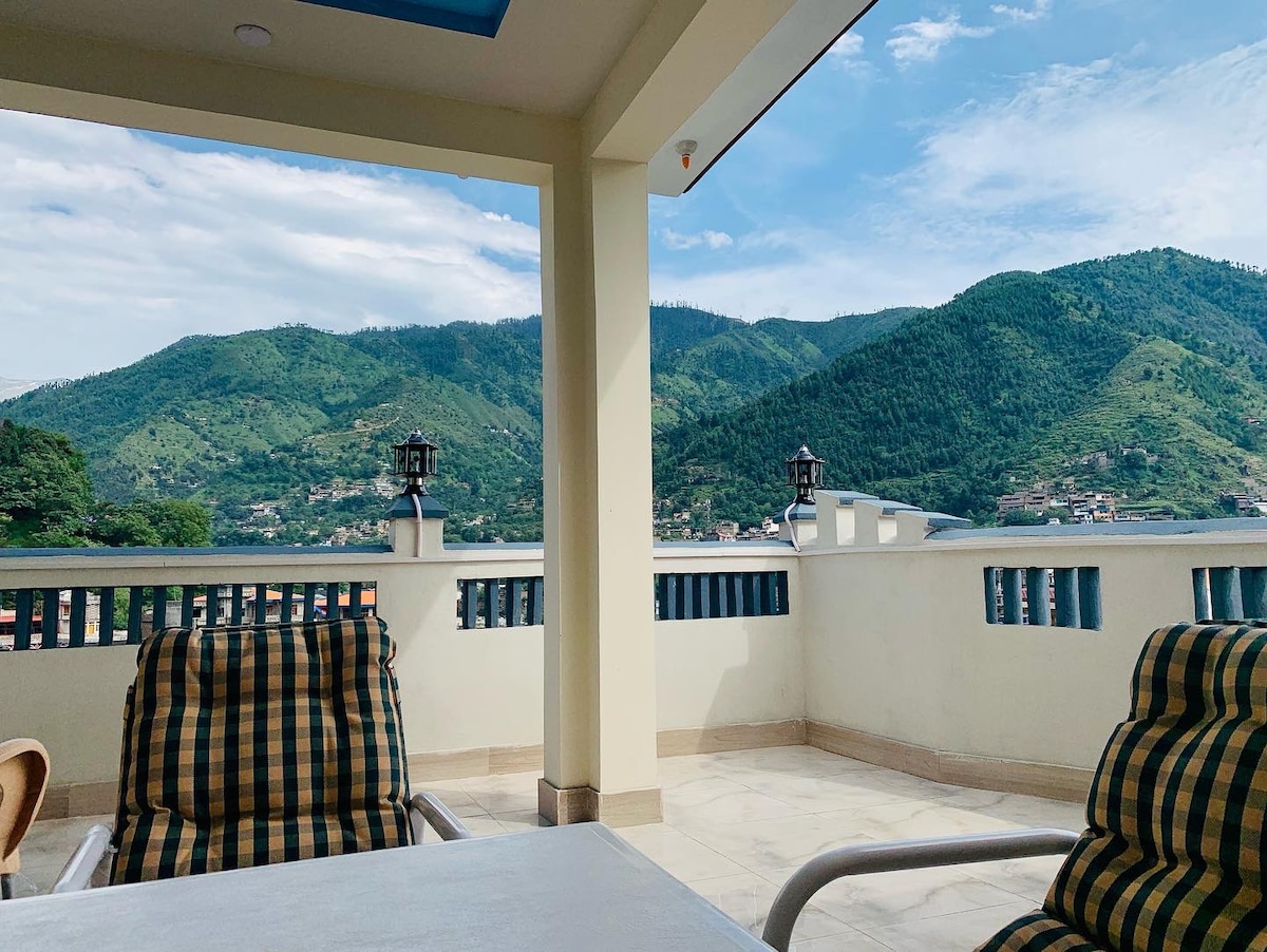 A spacious terrace is presented with comfortable seating options, featuring checkered patterns. The backdrop offers a panoramic view of verdant hills under a bright sky, enhancing the serene outdoor environment. A table is positioned centrally, inviting outdoor dining experiences.