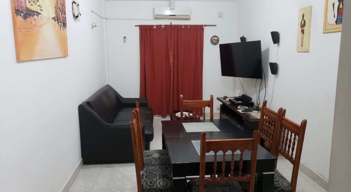A living area is seen, featuring a dark sofa positioned beside a wooden dining table surrounded by chairs. A flat-screen television is mounted on the wall opposite the seating area. Red curtains frame a window, while light tiles cover the floor.