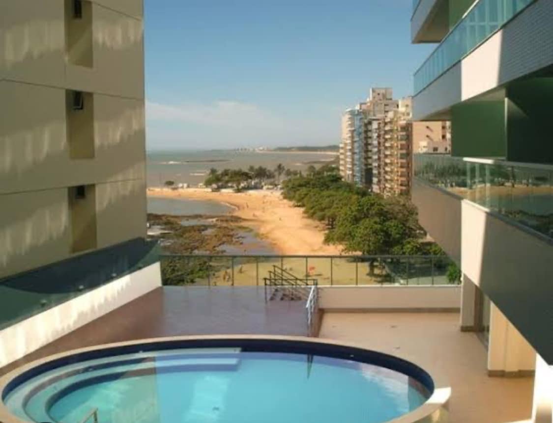 A view from a balcony showcases a circular pool overlooking the beach and ocean. The sandy shore is framed by lush greenery and lined with buildings in the background, under a clear sky with soft clouds.