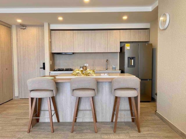 A modern kitchen area features light wooden cabinetry and a countertop with three upholstered stools. Stainless steel appliances, including a refrigerator and gas stove, are visible. Warm lighting illuminates the space, creating a functional area for meal preparation and dining.