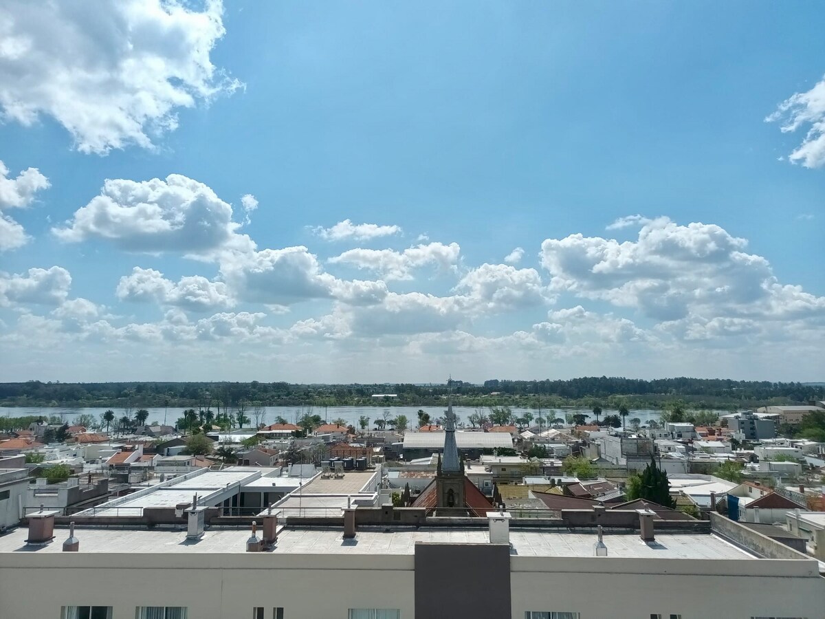 A panoramic view showcases a blend of urban and natural landscapes. The image captures rooftops of buildings, with a calm body of water and greenery in the background under a bright blue sky dotted with clouds.