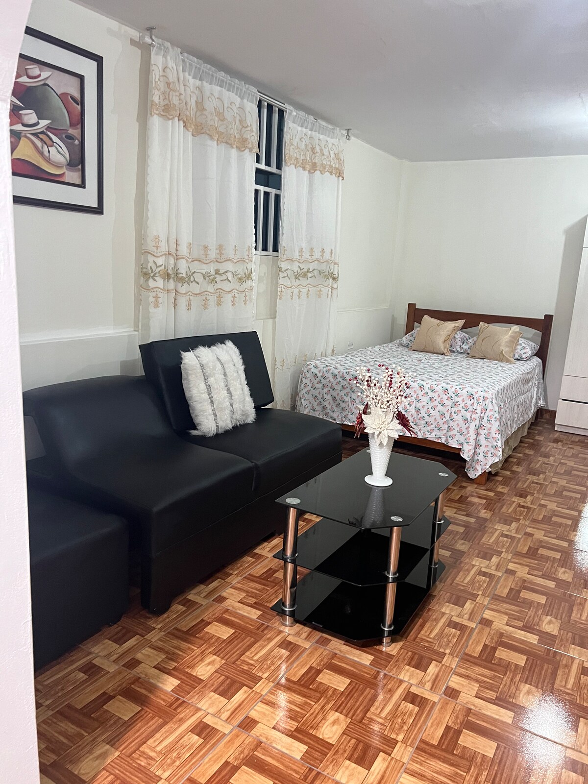 A compact living space features a black sofa with a decorative pillow and a glass coffee table at its center. A floral-patterned bed is situated nearby, flanked by two pillows. Light-colored curtains frame a window, illuminating the room's warm wooden flooring.