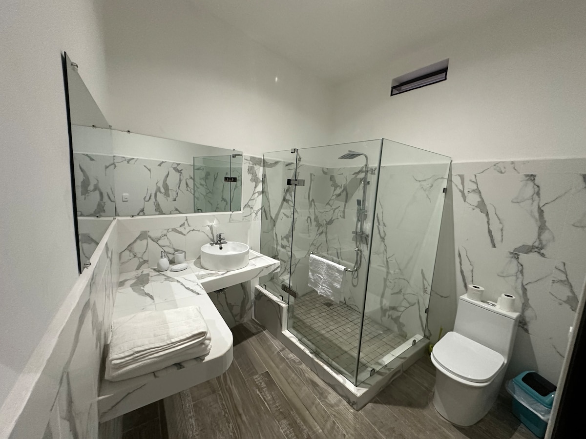 The bathroom features modern design elements, including a spacious glass shower enclosure and a floating vanity with a sink. Light-colored marble walls enhance the space, while a large mirror reflects the natural light coming in. A toilet is positioned adjacent to the shower.