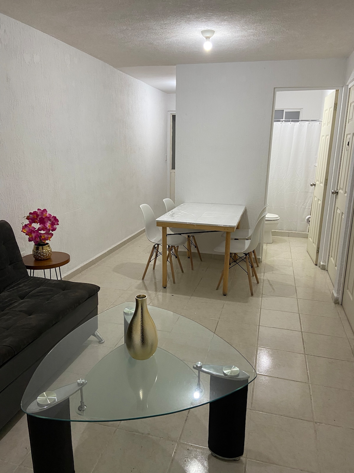 A dining area features a light wooden table surrounded by white chairs. A modern glass table sits in the foreground, while a black sofa and a decorative vase rest nearby. Soft lighting highlights the clean, neutral walls of the space.