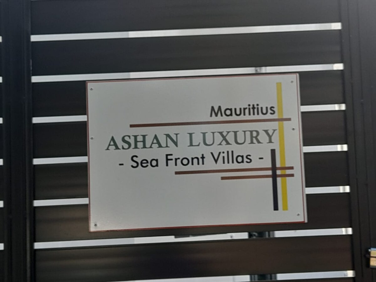 The entrance sign features the text 'Ashan Luxury - Sea Front Villas' prominently displayed. The sign is bordered in neutral tones, with stripes in yellow and black adding a contemporary touch. It is mounted on a sleek, dark panel, indicating the property's name and location in Mauritius.